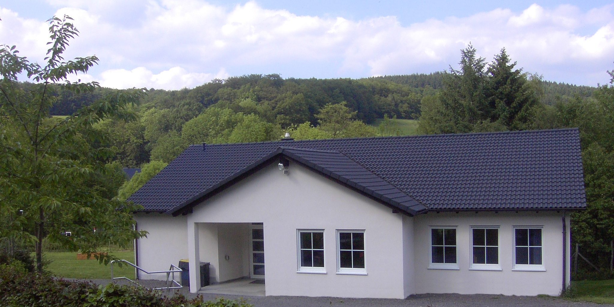 Gemeindehaus der LKG Vollme: weißes Gebäude mit dunklem Dach und überdachtem Eingang, im Hintergrund bewaldete Hügel